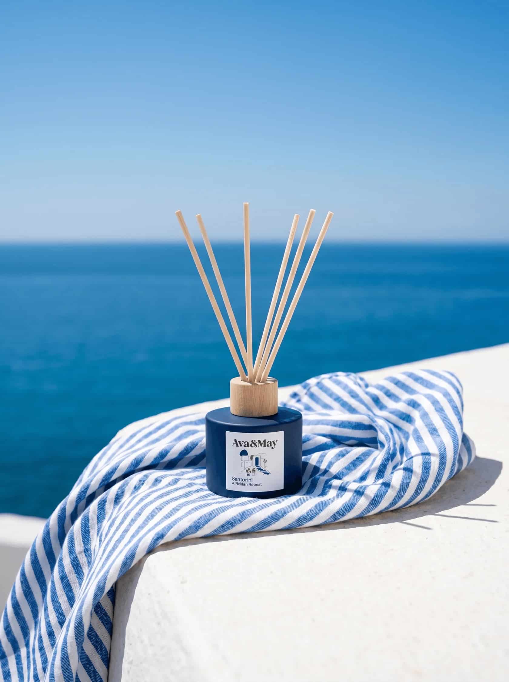 Ava&May Santorini Duftstäbchen auf einer weißen Mauerkante mit blau-weiß gestreiftem Tuch, im unscharfen Hintergrund das tiefblaue Ägäische Meer unter wolkenlosem Himmel. Das Bild fängt das griechische Inselgefühl ein, das viele Nutzer in ihren Santorini Ava&May Erfahrungen 2026 mit dem Duft nach Zeder, Rosmarin und Tonkabohne verbinden, ein Stück Urlaub für Zuhause. #santorini #griechenland #urlaubsfeeling #avamay #meerblick