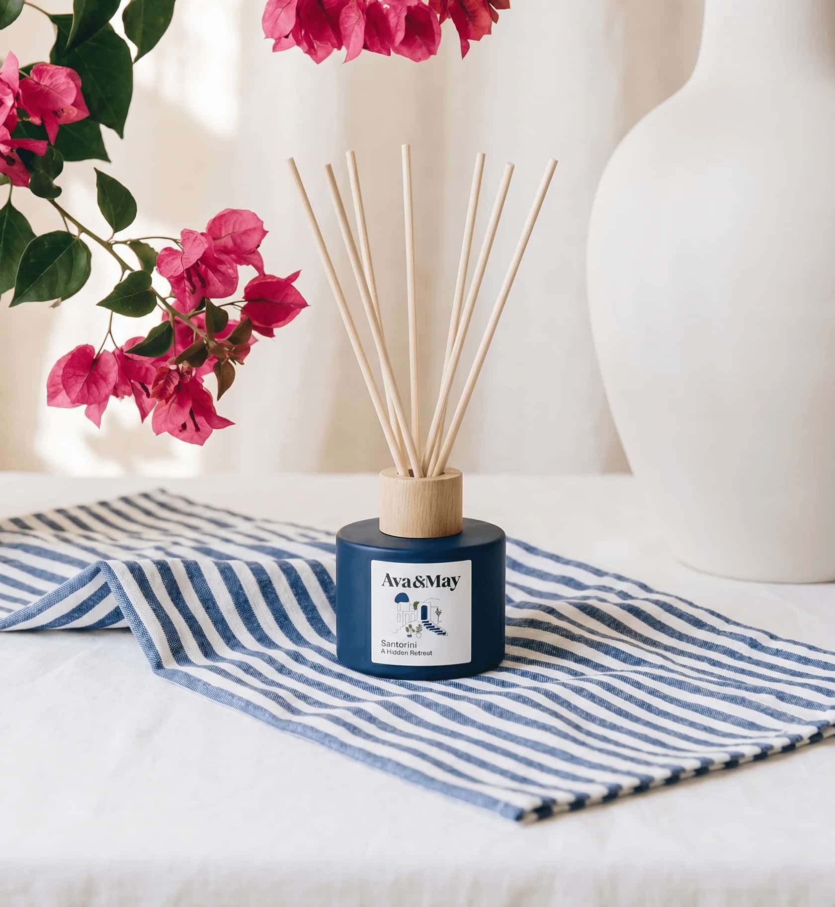 Ava&May Santorini Raumdiffuser in dunkelblauer Flasche mit Rattan-Stäbchen, präsentiert auf einem blau-weiß gestreiften Leinentuch. Im Hintergrund hängt ein Zweig pinker Bougainvillea ins Bild, daneben eine schlichte weiße Keramikvase. Erfahrungsberichte 2026 beschreiben den Duft als warm-herb mit holzigem Tonkabohnen-Unterton, ideal für Wohn- und Schlafzimmer. #avamay #santorinifeeling #bougainvillea #homefragrance #mediterrandeko