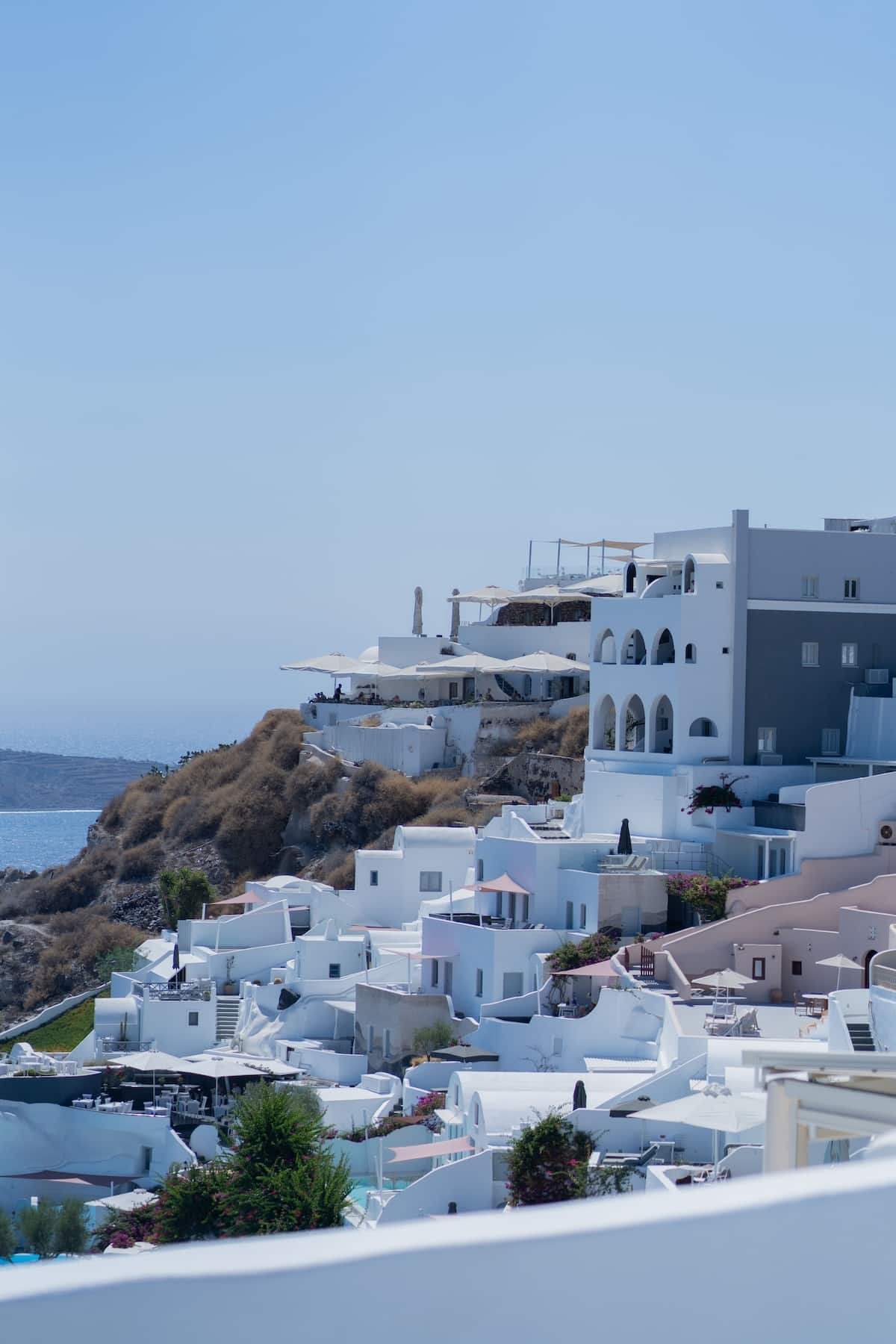 Verschachtelte weiße Häuser mit Bogengängen, Sonnenschirmen und Terrassen an der Steilküste von Santorini, organisch in den Hang integriert. Im Hintergrund das offene Meer, karge mediterrane Hügelvegetation und klarer blauer Himmel. Genau diese Atmosphäre greift der Ava&May Duft mit Zeder, Rosmarin und Tonkabohne auf. #santorini #kykladenarchitektur #griechenland #ägäis #inselurlaub